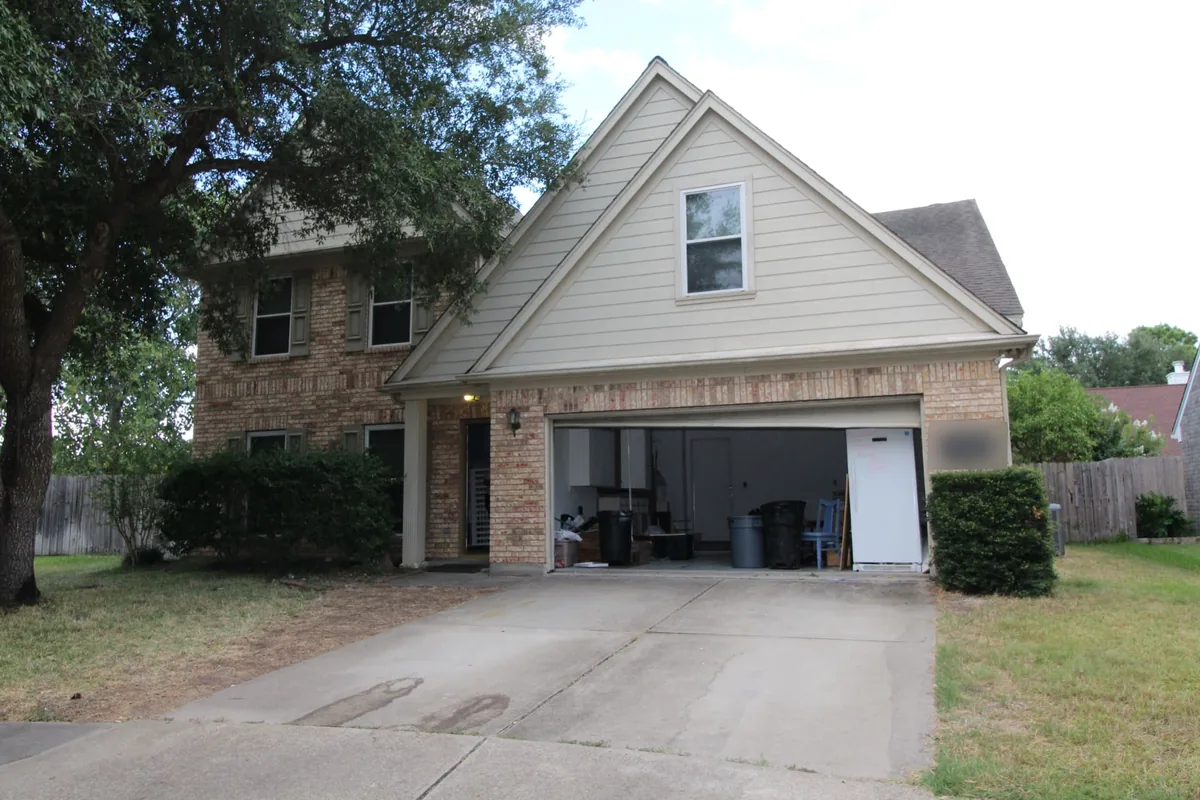 Two-story brick house with light gable siding, an open garage, concrete driveway and a large shade tree; house number blurred
